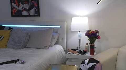 Media: Video of a modern bedroom with a white bed, white headboard, and a bedside table with a lamp and flowers. Background includes a framed art piece and scattered makeup items.