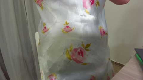 Media: A video of a person wearing a white shirt with large pink and yellow rose patterns. The background features a beige wall and a partially visible piece of furniture.