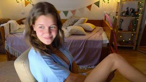 Media: A video of a young woman with shoulder-length brown hair, wearing a light blue shirt, sitting in a cozy bedroom with a wooden bed, fairy lights, and a bookshelf.