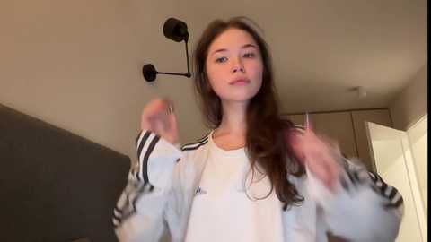 Media: Video of a young Caucasian woman with long brown hair, wearing a white Adidas jacket and a white top, standing in a modern bedroom with beige walls and a black wall.