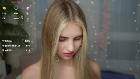 Media: A video of a young, fair-skinned, blonde woman with straight hair, wearing a strapless top, standing indoors with a blurred background.