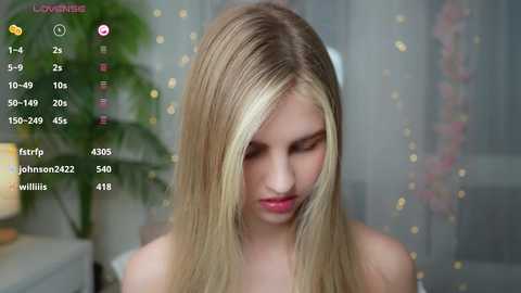 Media: Video of a young, light-skinned blonde woman with straight hair, wearing a pink top, in a softly lit room with blurred background and digital camera controls.