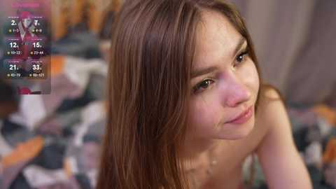 Media: Video of a young, fair-skinned woman with long, straight brown hair, wearing a strapless top, sitting on a bed with a colorful, patterned blanket. Background is blurred.