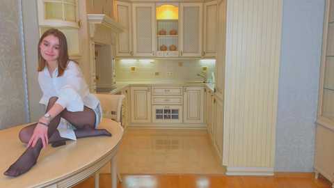 Media: Video of a young Caucasian woman with light brown hair, sitting on a round wooden table in a light-colored, modern kitchen with white cabinets and wooden floors. She wears a white blouse, black tights, and a playful smile.