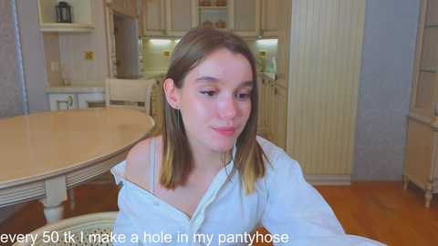 Media: Video of a young woman with shoulder-length brown hair, wearing an off-shoulder white shirt, sitting in a kitchen with beige cabinets and a round table. Text reads: \"eveny 50 tk make a hole in my pantyhose.\