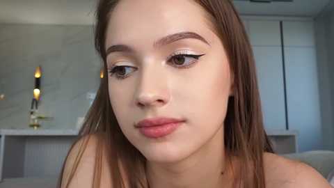 Media: Video of a young woman with fair skin, brown eyes, and straight brown hair, wearing natural makeup and a soft pink lipstick. Background features a modern living room with gray walls, a white shelf, and a lamp.