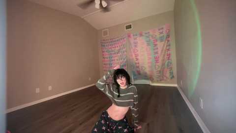 Media: Video of a young woman with a slim build, wearing a green striped crop top and floral pants, posing playfully in a dimly lit room with a tie-dye wall hanging.