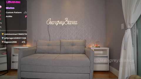 Media: Video of a modern, minimalist bedroom with a light gray tufted sofa, white bedside tables, and a soft gray textured wall featuring \"JeremyCam\" in cursive.