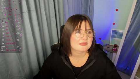 Media: Video of a middle-aged woman with shoulder-length brown hair, wearing glasses, a black hoodie, and a necklace, seated in a room with light blue curtains and a framed wall chart.