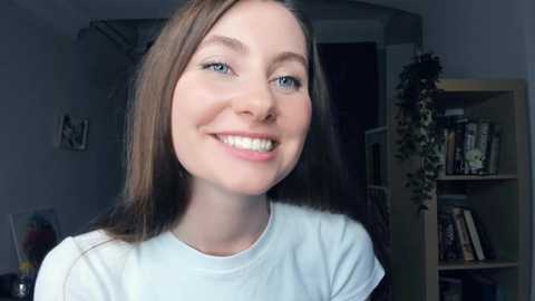 Media: Video of a smiling young woman with straight, long brown hair, light skin, and blue eyes, wearing a white t-shirt, standing in a dimly lit room with a bookshelf and plants in the background.