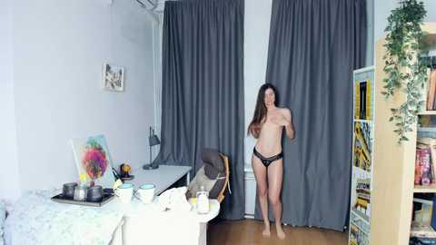 Media: A video of a topless, slender woman with long brown hair, wearing only black lace panties, standing in a modern, well-lit bedroom with grey curtains, a desk, and a bookshelf.