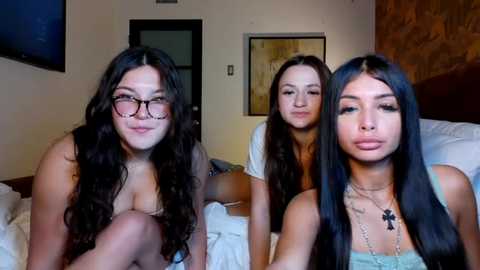 Media: Video of three young women, one wearing glasses, sitting on a bed in a dimly-lit bedroom with beige walls, a wooden headboard, and framed art.