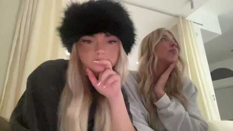 Media: Video of two blonde women in a bedroom; one wears a black fur hat, the other a gray hoodie, both looking contemplative.