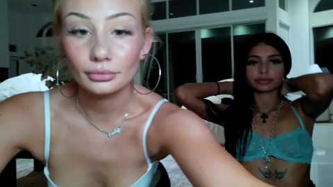 Media: Video of two young women with light skin and platinum blonde hair, one wearing a light blue bra, the other a turquoise bra, posing indoors with white bedding and a mirror in the background.