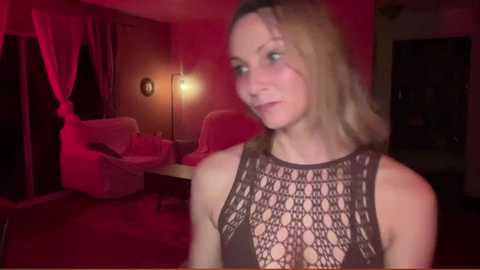 Media: Video of a fair-skinned woman with blonde hair in a brown, crochet-patterned top, standing in a dimly lit, cozy room with red lighting, soft furniture, and a lamp.