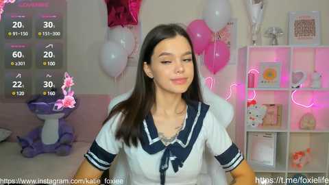 Media: Video of a young woman with long dark hair, wearing a white sailor shirt, smiling in a cozy, pink-themed room with balloons, plush toys, and shelves.