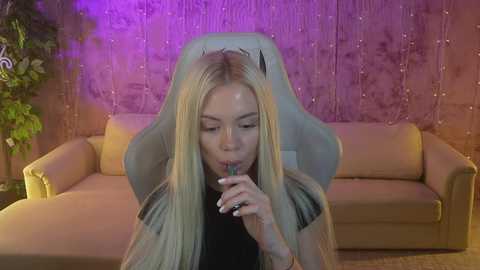 Media: Video of a blonde woman in a gaming chair, eating ice cream with a lollipop, in a cozy living room with a beige couch and purple string lights.