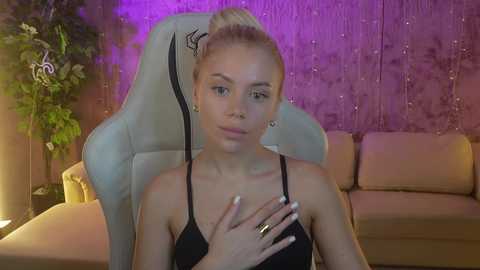 Media: Video of a blonde, fair-skinned woman with a slender physique, wearing a black spaghetti-strap top, seated on a white gaming chair with a plant to the left and a beige couch in the background.