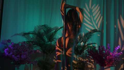 Media: Video of a nude woman with long, wavy hair, holding a pole, in a dimly lit jungle-themed room with green walls, ferns, and purple orchids.