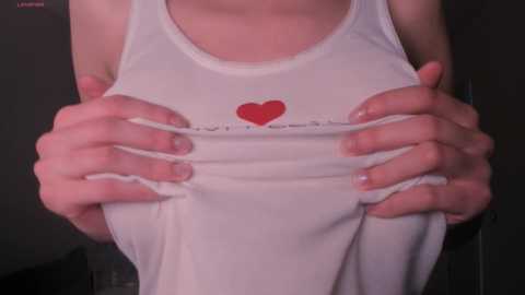 Media: Video of a light-skinned woman with small breasts wearing a white tank top, hands lifting the top to reveal a red heart on her chest, against a dark background.