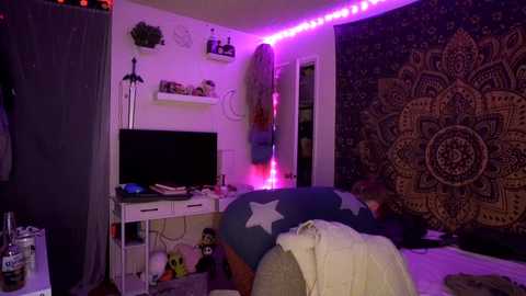 Media: Video of a dimly lit, eclectic bedroom with purple LED strip lighting, white shelves, a TV, a large mandala tapestry, and a Superman comforter.