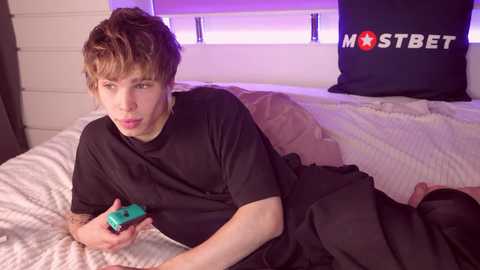 Media: Video of a young person with messy brown hair, wearing a black t-shirt, lying on a bed with a white blanket, holding a green Nintendo Switch, in a dimly lit room.