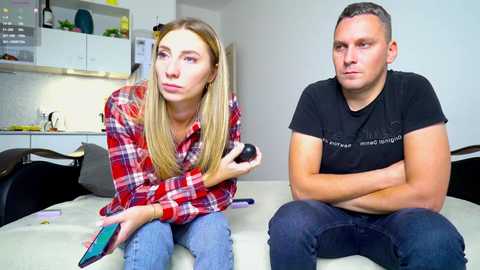 Media: Video of a young blonde woman in a red plaid shirt, holding a black smartphone, sitting next to a man in a black T-shirt, arms crossed, in a modern kitchen with white cabinets and stainless steel appliances.