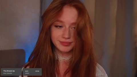 Media: Video of a young red-haired woman with pale skin, wearing a white lace dress, smiling slightly. Background features blurred beige and blue tones.