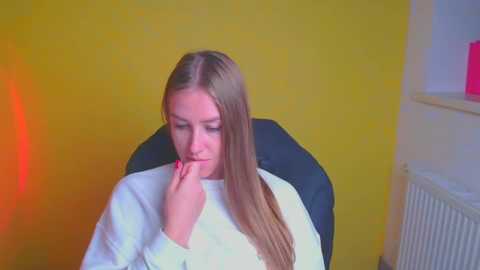 Media: Video of a young woman with long brown hair, wearing a white sweater, sitting in a black chair against a yellow wall. She has a contemplative expression, with her hand to her mouth.