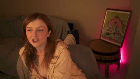 Media: Video of a young woman with long, auburn hair, wearing a beige shirt, sitting on a gray couch in a dimly lit room with a framed painting on a wooden side table.