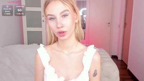 Media: Video of a young woman with fair skin, straight blonde hair, and blue eyes, wearing a white frilled top. She has a nose piercing and a small tattoo on her left arm. The background is a modern bedroom with light pink accents.