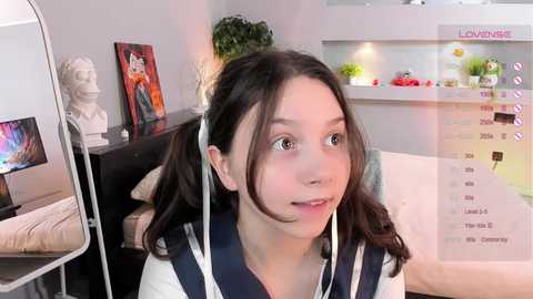 Media: Video of a young Asian woman with long black hair in pigtails, wearing a navy blue and white sailor dress, sitting in a modern living room with a calendar, plants, and art on the wall.