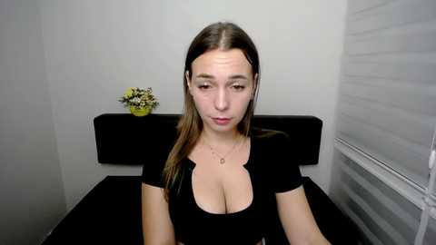 Media: Video of a young Caucasian woman with light skin, long brown hair, and large breasts, wearing a black top, sitting in a minimalist, white-walled room with a floral arrangement on a black shelf.