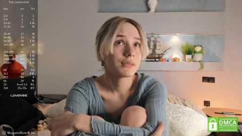 Media: Video of a young woman with short blonde hair, wearing a blue top, cradling a baby, in a cozy bedroom with a nautical-themed shelf and a modern lamp.