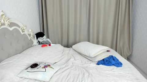 Media: Video of a tidy, modern bedroom with a white metal headboard, beige curtains, and a plush white bed featuring a stuffed animal, folded clothes, and an open book.