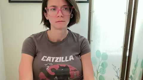 Media: Video of a young woman with short brown hair, wearing pink glasses and a grey t-shirt with a \"Catspaw\" design. She stands in a bathroom with a floral shower curtain.