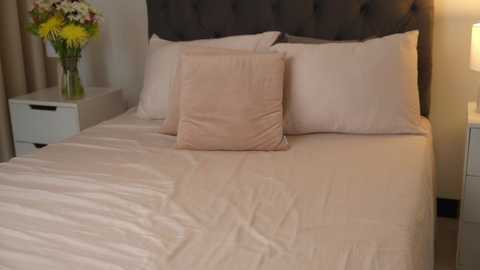 Media: Video of a neatly made bed with a tufted dark gray headboard, white pillows, and a beige quilted pillow. A vase with yellow flowers sits on a white nightstand to the left.