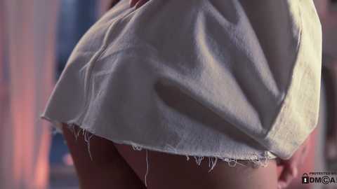 Media: A close-up video of a woman's lower back and buttocks, wearing a frayed, white, short-sleeved dress with visible skin underneath. The background is blurred, featuring pastel hues and vertical stripes.