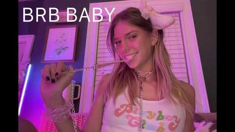 Media: Video of a young woman with long blonde hair, wearing a white tank top with colorful text, a choker, and a pink bunny ear headband. She holds a whip and smiles. Background includes framed art and a window. Text reads \"BBB BABY\" in white.