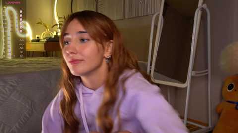 Media: Video of a young woman with long auburn hair, wearing a lavender hoodie, sitting in a cozy bedroom with a gray bed, a white ladder, and a plush toy.