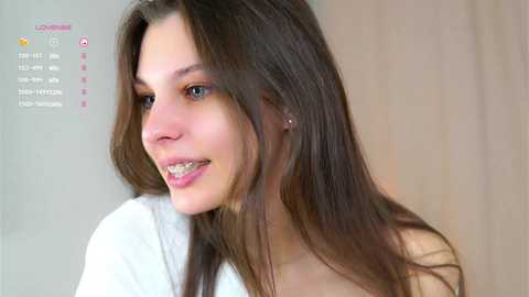 Media: Video of a young Caucasian woman with long brown hair and braces, smiling, wearing a white top. Background is blurred beige.