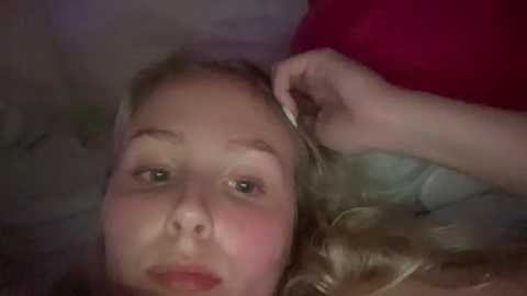 Media: Video of a blonde girl lying on a bed, her fair skin and rosy cheeks visible, with a hand gently touching her hair.