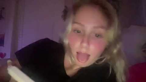 Media: A video of a young woman with light skin, blonde hair, and a black T-shirt, joyfully sticking out her tongue, in a dimly lit bedroom.