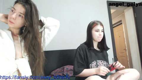 Media: Video of a young woman with long, straight dark hair, wearing a white shirt, posing provocatively; another young woman with black hair, wearing a black \"NOFX\" t-shirt, sitting on a bed, holding a red vibrator.