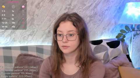 Media: Video of a young woman with fair skin, glasses, and long brown hair, sitting on a checkered sofa, wearing a brown sweater. Background features a gray wall with a blue lamp and plant.