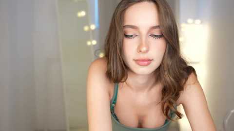 Media: Video of a young Caucasian woman with long brown hair, fair skin, and green eyes, wearing a low-cut green top, looking down, in a softly lit room with white curtains and fairy lights.