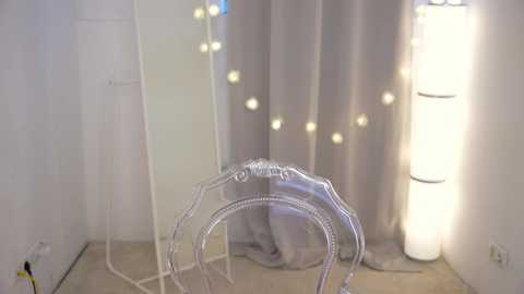 Media: A video of a modern, minimalist room with white walls and a light wood floor. A silver, wire-framed chair stands in the center, with a white mirror and a string of fairy lights in the background.