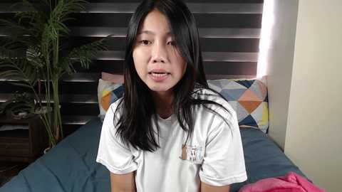 Media: Video of an East Asian woman with straight black hair, wearing a white T-shirt, sitting on a bed with a blue blanket and a colorful geometric-patterned pillow. Background includes a dark wooden headboard and a green plant on a nightstand.