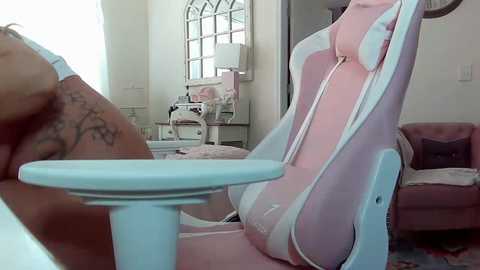 Media: Video of a plush, pink gaming chair with white accents and a small table in a brightly lit, modern living room with a large window and soft furnishings.