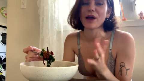 Media: Video of a fair-skinned woman with short brown hair, wearing a light gray lace bra, holding a salad bowl with a fork, sitting in a kitchen with white walls and a window.